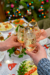 Human hands holding glasses with sparkling wine. Friends or family toasting with champagne against festive Christmas table and decorated new year tree. Winter holidays celebration at cozy home