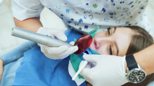 A dentist in white latex gloves uses a UV lamp with a red protective glass to solidify a dental filling. Dental restoration and material polymerization with UV light.