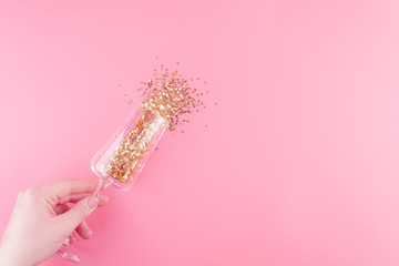 Female hand holds a glass of champagne filled with Christmas decorations in the form of gold stars on a pink pastel background.