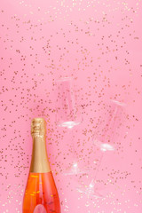 A bottle of pink champagne and an two empty glasses surrounded by gold confetti stars on a pink pastel background.