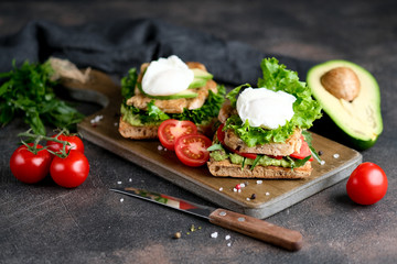 Delicious sandwich with avocado and poached egg, cherry tomatos and arugula on a dark background.