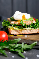Delicious sandwich with avocado and poached egg, cherry tomatos and arugula on a dark background.
