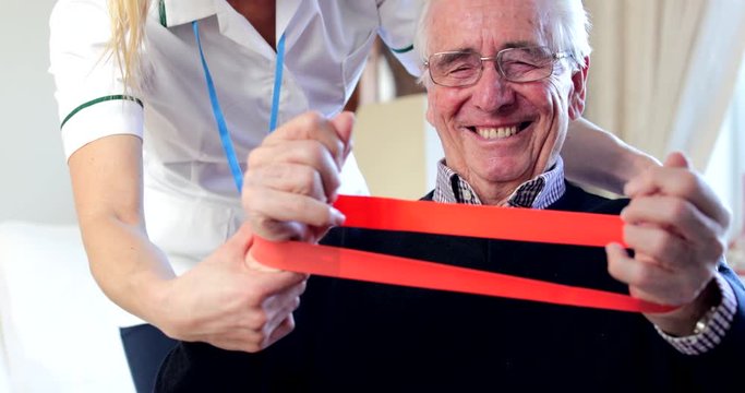 Female Physiotherapist Helping Senior Man To Use Resistance Band At Home