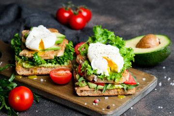 Delicious sandwich with avocado and poached egg, cherry tomatos and arugula on a dark background.