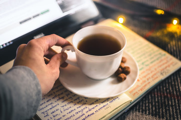 Cup of hot tea with a hand in a Christmas atmosphere with a laptop, a cozy blanket and a book in warm colors