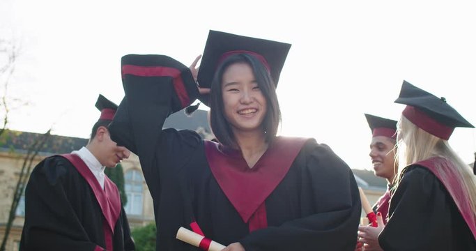 Portrait of the young beautiful Asian girl, master student graduate in the academic outfit and with diploma in hands smilingto the camera and then tossing up in the air her cap. Outdoor.