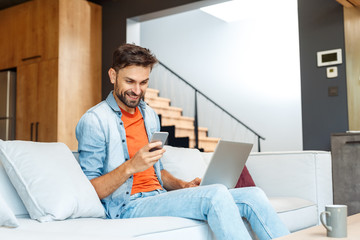Young adult man sitting at home, using smartphone and laptop