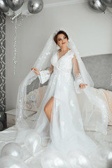 Young woman in wedding robe in luxury interior with a mass of white and grey balloons. Charming young bride with make up and hair style. Morning of the bride preparations. Girl standing on the bed