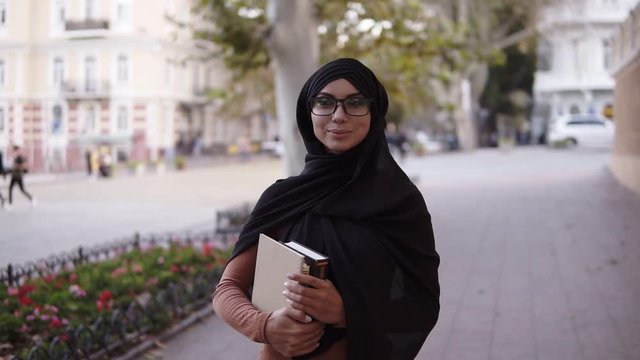 Confident attractive muslim woman in hijab and glasses walking by streetclose to the college or university, beautiful young female in black clother, carrying books in her arms