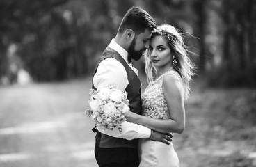 Sensual Black and white foto of bride and groom. Stylish couple of happy newlyweds posing in the park on their wedding day. Handsome bearded groom admires and kisses pretty bride. Together.