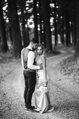 Fototapeta premium Sensual Black and white foto of bride and groom. Stylish couple of happy newlyweds posing in the park on their wedding day. Handsome bearded groom admires and kisses pretty bride. Together.
