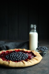 Homemade backing. Delicious galette with plums and blueberries with cup of milk on a dark background copy space.