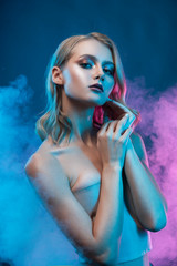 Beautiful blonde young girl with trendy makeup touches her face and hair with her hands, surrounded with blue and violet light in theatrical smoke. Clear, healthy skin. Advertising, commercial design.