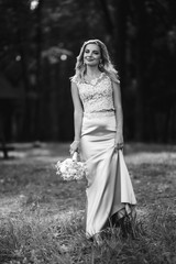 Black and white foto of beautiful bride with a wedding bouquet for a walk in the park. Happy newlywed woman stands among green bushes in the garden. Smiling bride. Wedding day. Fashion bride.