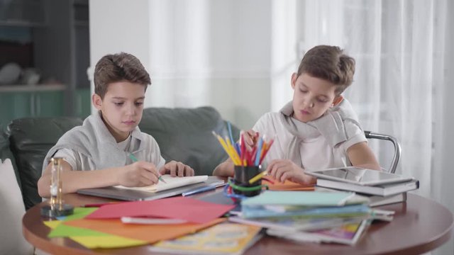Two creative Caucasian twin brothers sitting at the table and drawing with colorful pencils. Cute boy looking at his bro's picture and showing thumb up. Siblings doing homework together. Education.