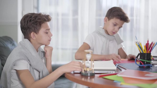 Side view of brunette Caucasian boy turning sandglass and his twin brother starting drawing at the background. Siblings competing in speed. Generation Z doing homework together.