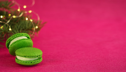 Green macaron with fondant on red paper background. Near a branch of a Christmas tree with a garland. Close-up, copy space for text. Postcard or banner concept.
