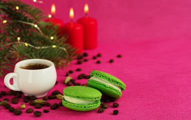 Green macaron with fondant on a red paper background. Nearby is a cup of espresso, roasted coffee grains. Christmas tree branch with a garland and burning candles. Close-up, copy space for text.