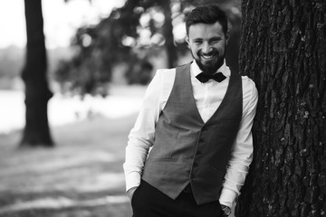 Black and white portrait of confident handsome man in black suit with bowtie posing in the park. Luxury groom in suit. Businessman. Happy smiling groom newlywed. Rich man at wedding day. 
