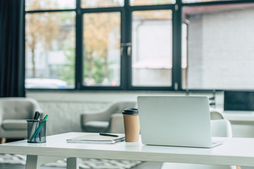 laptop, disposable cup, notebook and pen holder on white table in modern office