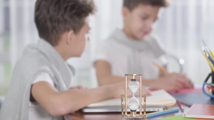Close-up of turned sandglass staying at the table as two blurred schoolboys drawing or writing at the background. Twin brothers in similar clothes doing homework and competing in speed. Education.