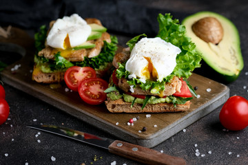 Delicious sandwich with avocado and poached egg, with green leaves and tomatoes on a dark background. Healthy breakfast.