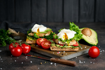 Delicious sandwich with avocado and poached egg, with green leaves and tomatoes on a dark background. Healthy breakfast.
