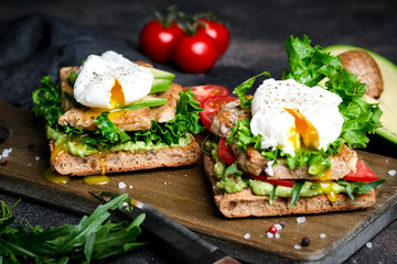 Delicious sandwich with avocado and poached egg, with green leaves and tomatoes on a dark background. Healthy breakfast.