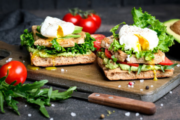 Fototapeta na wymiar Delicious sandwich with avocado and poached egg, with green leaves and tomatoes on a dark background. Healthy breakfast.