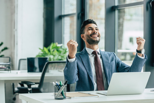 happy businessman looking up and showing winner gesture while sitting at workplace - Powered by Adobe