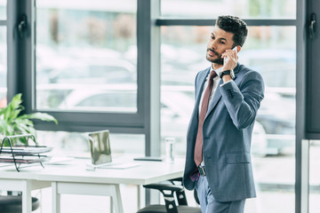 serious businessman standing near desk and talking on smartphone