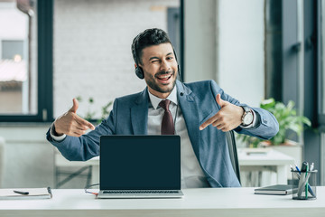 cheerful call center operator winking at camera and pointing with fingers at laptop with blank...