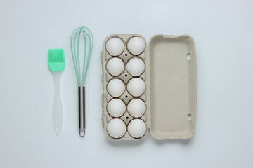 The concept of cooking. Cardboard tray of eggs, kitchen tools (whisk, brush) on white background. Top view