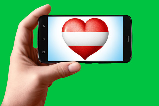 Austria flag in the shape of a heart on the phone screen. Smartphone in hand shows the heart of the flag on the background hromakey.