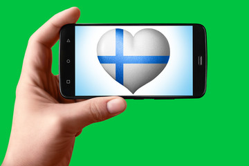 Finland flag in heart shape on the screen. Smartphone in hand shows the heart of the flag on the background hromakey.