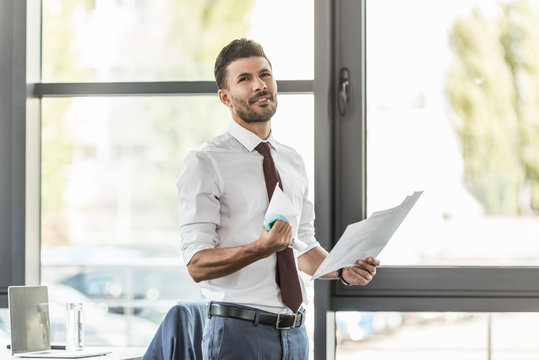 upset businessman holding documents and crumpling paper while looking away in office