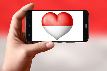 Monaco flag in the shape of a heart on the phone screen. Smartphone in hand shows a heart flag.