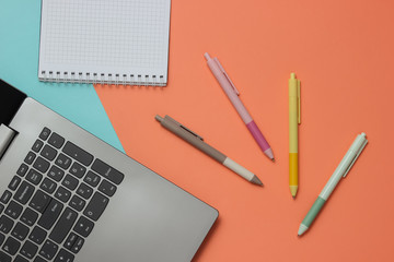 Laptop and many colored pens with notebook close-up on blue pink background. Top view