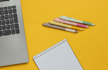 Laptop and many colored pens with notebook close-up on yellow background. Top view