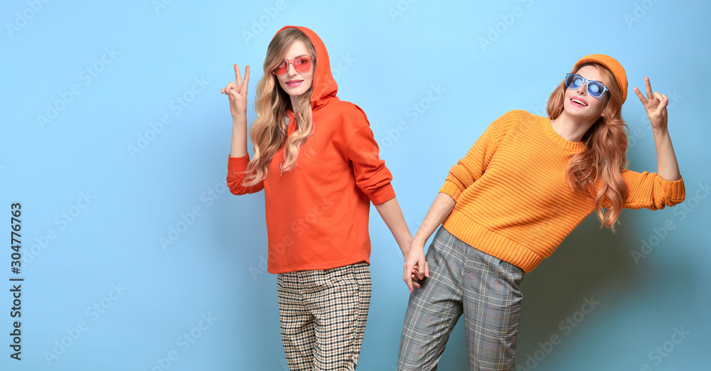 Wall mural Fashionable beautiful woman in stylish outfit, makeup, fun shows peace sign. Two happy blonde redhead girl, trendy orange jumper, hoody, fashion hair. Cheerful sister friend, funny colorful banner