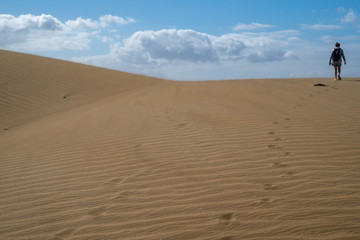 Fototapeta na wymiar Person is walking on sand dunes during sunny and windy day in the Natural Reserve of Dunes of Maspaloma in Gran Canaria with sand dust and foot steps. Canary Islands, Spain