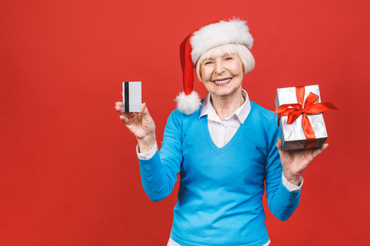 Happy Mature Woman With A Gift And Credit Card. Isolated Over Red Background. Beautiful Stylish Senior Lady Opening Her Christmas Gift. Elegant Mature Woman Holding A Red Gift Box.