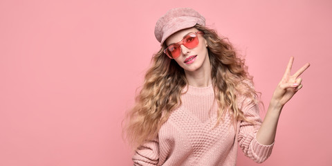 Easy-going Woman happy smiling in Stylish fashion pink colored outfit. Beautiful young Girl in Trendy jumper, make up. Joyful blonde lady shows peace sign in pink cap, fashionable happiness concept