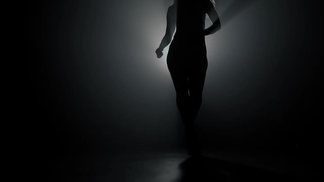 Silhouette of beautiful dancer on smoky dark background.Spotlight shines back against sexy girl. She moving seductively.