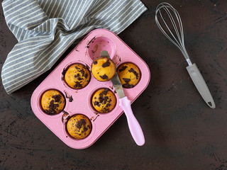 Cupcakes in pink cupcake mold, grey striped towel, napkin, kitchen spatula with pink handle on dark background.