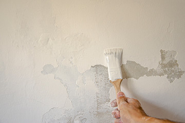 Hand with a brush in white paint on a white wall background with peeling paint - repair and painting the walls