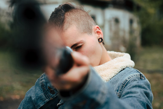 Young woman with short hair and a punk style, holds an old double-barrel shotgun in an abandoned estate. She is pointing at the camera.