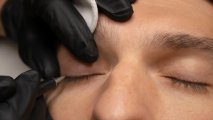 Young attractive man eye close-up with the master hand in black gloves and permanent makeup machine with a needle. Male permanent makeup procedure.