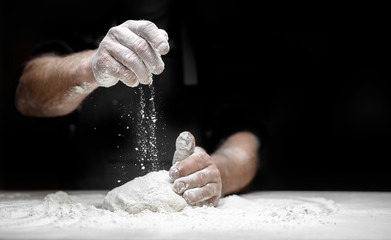 White flour flies in air on black background, pastry chef claps hands and prepares yeast dough for...