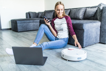 Young woman sitting on the floor with laptop and control robot vacuum cleaner from the phone